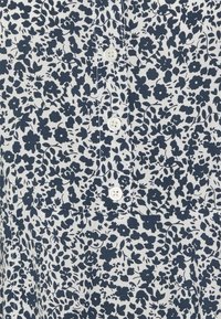 Vitt tyg med täta små mörkblå blommönster och fyra synliga vita knappar ner i mitten.