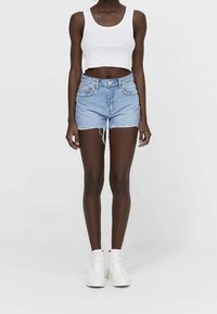 Mujer vestida con una camiseta blanca de tirantes corta, pantalones cortos de mezclilla de cintura alta en un tono azul claro con bordes deshilachados, y zapatillas altas blancas, de pie.