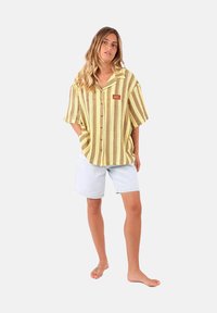 Chemise à manches courtes, boutonnée, en rayures jaunes et brunes, avec un col pointu et un petit écusson sur la poitrine. Associée à un short en denim clair.