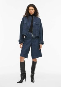 Džinsa jaka ar priekšējām kabatām, īss griezums un uzlocītas piedurknes, kopā ar atbilstošiem džinsu šortiem un plata josta. Augsti melni zābaki ar papēdi.