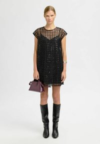 Moteris stovi vilkėdama juodą permatomą suknelę su blizgučiais, juodus iki kelių batus, sidabrinę apyrankę, laikydama mažą tamsiai violetinę rankinę.