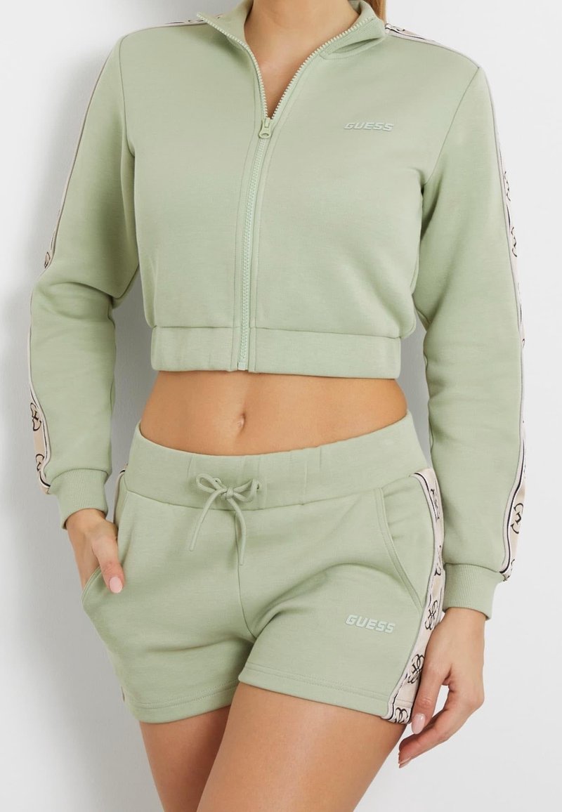 Femme portant une veste courte zippée vert clair et un short assorti avec cordon de serrage et détails du logo sur les manches et les côtés.