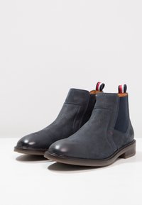 Tmavě modré kožené kotníkové boty s elastickými bočními panely, zaoblenými špičkami a kontrastními červenými a bílými poutky na zadní straně.