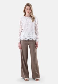 Weißes Spitzenoberteil mit langen Ärmeln und floralem Muster, kombiniert mit lockeren hellbraunen Hosen und beigen geflochtenen Sandalen.