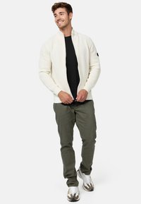 Hvid, ribbet strikket zip-up jakke båret over en sort skjorte, parret med olivengrønne bukser og hvide sneakers, med et logo på ærmet.