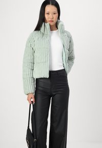 Veste courte matelassée vert clair avec texture en côtes, fermeture éclair intégrale et col haut. Associée à un pantalon en simili cuir noir et un petit sac noir.