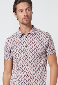 Camisa de botones de manga corta con un patrón geométrico en rojo, blanco y azul claro sobre un fondo crema. Fabricada con un material ligero.
