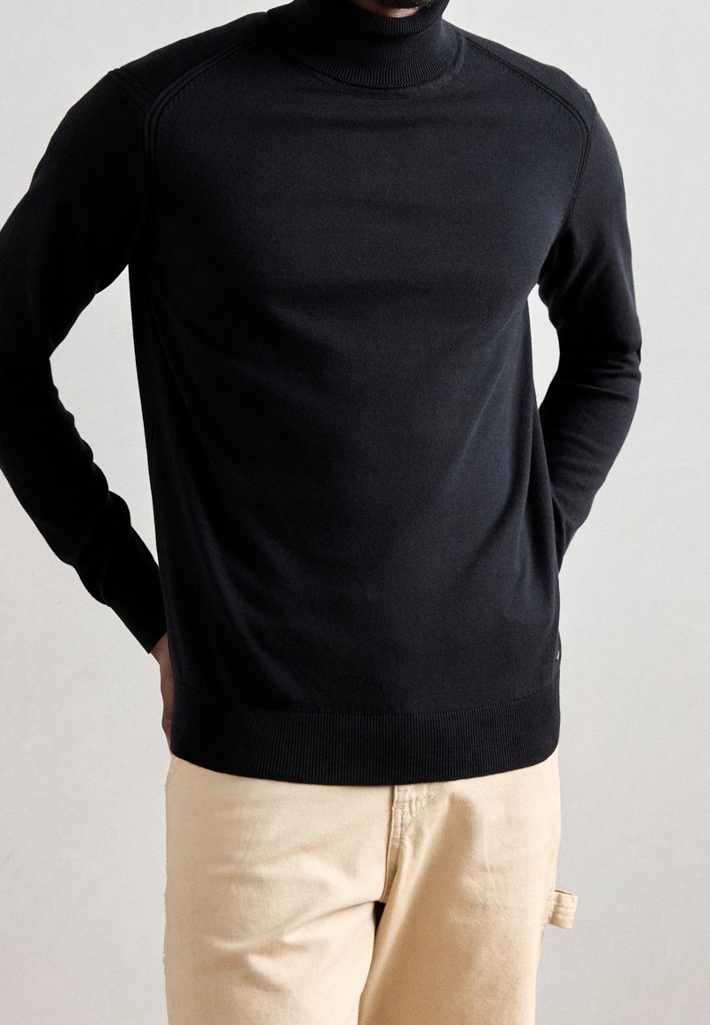 Pull à col roulé noir à manches longues avec poignets côtelés. Design lisse et ajusté avec un léger éclat, porté avec un pantalon de couleur claire.