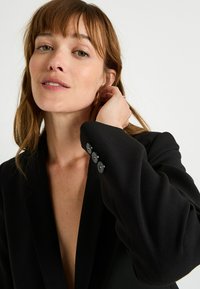 Blazer noir avec un décolleté en V profond, coupe ajustée et quatre boutons sur les manches, fabriqué à partir d'un tissu lisse et texturé.