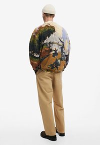 Maglione lavorato a maglia con un motivo paesaggistico colorato, scollo rotondo e vestibilità morbida, abbinato a pantaloni beige e scarpe nere.