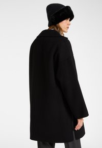 Abrigo de mezcla de lana negro con corte holgado, gran solapa y silueta recta. Lleva puesto un gorro de punto negro de canalé con una banda de piel sintética.