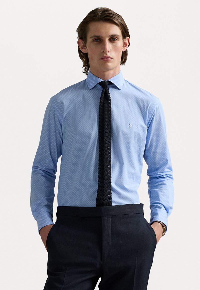 Chemise bleu clair à manches longues avec un motif discret à pois, associée à une cravate noire texturée. Portée avec un pantalon sombre et une montre.