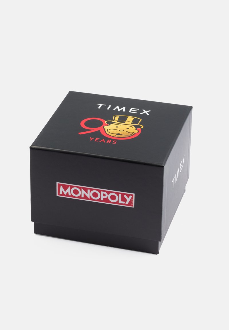 Caja de cartón negro con los logotipos "TIMEX" y "MONOPOLY", con un personaje dorado de Monopoly y "90 AÑOS" en rojo. Diseño elegante, forma cuadrada.