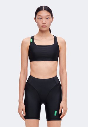 Jeune femme aux cheveux noirs et raides porte un soutien-gorge de sport noir et un short cycliste taille haute assorti avec des étiquettes vertes sur un fond uni.