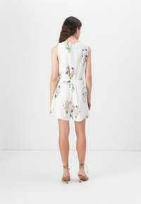 Jumpsuit senza maniche bianco con motivo floreale in vari colori. Presenta una vita stretta e pantaloni corti, abbinato a tacchi a spillo marroni.