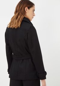 Abrigo negro con cinturón, de diseño ajustado, cuello amplio y puños doblados. La tela parece suave con costuras estructuradas y detalles en la parte trasera.