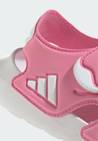 Różowe sportowe sandały z teksturowaną gumową cholewką, białymi akcentami i detalem w postaci logo z trzema paskami. Projekt wyposażony w zapięcie na pasek zapewniające bezpieczeństwo.