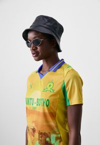 Maglietta da calcio gialla con accenti verdi e "HUNTU-BOTHO" in grassetto verde. Accompagnata da un cappello a secchiello nero e occhiali da sole argentati.