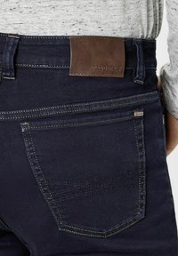 Dark denim jeans with a brown leather back patch, visible stitching, and a pocket design. The material appears textured and durable.