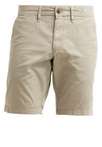 Short beige mi-longueur avec fermeture à bouton et zip, passants de ceinture et une petite étiquette de marque près de la taille.