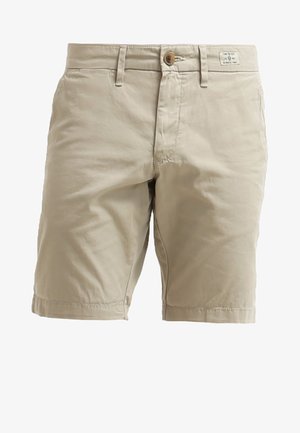 Mand iført hvid t-shirt, beige shorts og sorte bådskor, der holder solbriller, går mod en ensfarvet lys baggrund.