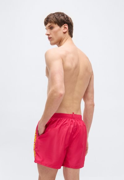 Άνδρας χωρίς μπλούζα με κόκκινο μαγιό με κίτρινο κείμενο στο πλάι, όρθιος με τα χέρια στις τσέπες, κοιτάζοντας πάνω από τον αριστερό του ώμο.