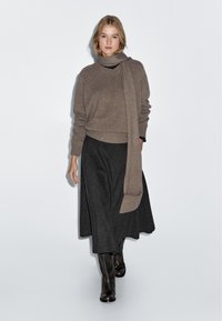 Brauner Strickpullover mit hohem Kragen und passendem Schal, kombiniert mit einem langen schwarzen gemusterten Rock und braunen kniehohen Stiefeln.