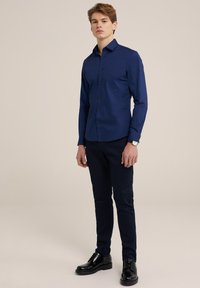 Cămașă albastru marin cu nasturi, dintr-un material neted, mâneci lungi, croială ajustată, cu guler clasic și nasturi închisi, asortată cu pantaloni închiși la culoare și pantofi negri.