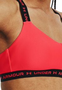 Brassière de sport rose avec une texture lisse, dotée de bretelles noires et d'une bande avec le logo "UNDER ARMOUR" en lettres rouges.