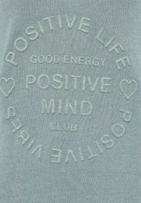 Hellblauer Stoff mit erhabener Stickerei, die den Text "POSITIVE LIFE GOOD ENERGY POSITIVE MIND CLUB" und Herzakzente zeigt.