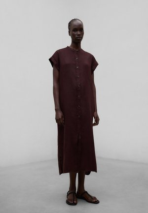 Mujer con vestido midi sin mangas, abotonado y de color marrón oscuro, y sandalias planas negras, de pie frente a un fondo liso de color gris claro.