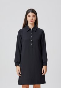 Robe noire à col, avec des manches longues et une patte de boutonnage. Fabriquée en tissu lisse, elle est dotée de cinq boutons ronds et de poignets fuselés.