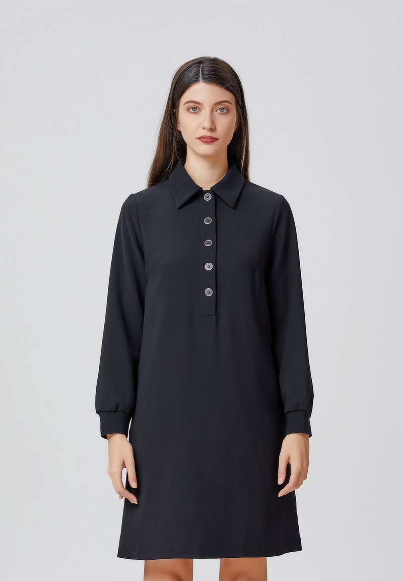 Robe noire à col, avec des manches longues et une patte de boutonnage. Fabriquée en tissu lisse, elle est dotée de cinq boutons ronds et de poignets fuselés.