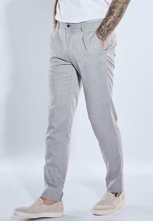 Mann trägt hellgraue Nadelstreifenhose, weiße Slip-On-Schuhe und weißes Hemd, Hände in den Taschen, sichtbare tätowierte Unterarme.