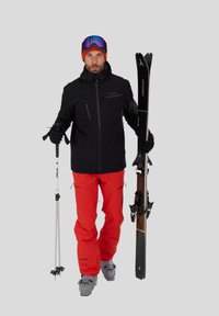 Lyžař v černé bundě a červených kalhotách, držící lyže a hůlky. Na hlavě má červenou čepici a mnohobarevné brýle, se šedými lyžařskými botami.