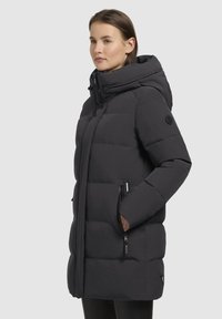 Giacca a vento nera con cappuccio, chiusura frontale con zip, pannelli cuciti, tasche laterali e patch con logo sulla manica. Materiale liscio e isolato.