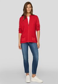 Roter gestrickter Cardigan mit Rippenstruktur, zwei Vorderentaschen und Reißverschluss. Trägt man über einem weißen Hemd, kombiniert mit blauen Jeans und Turnschuhen.