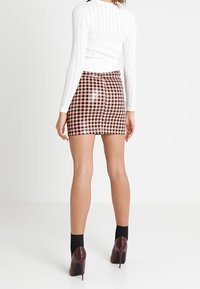 Czarno-brązowa mini spódniczka w kratę wykonana z błyszczącego materiału, zestawiona z białym, prążkowanym, długim topem z długim rękawem oraz czarnymi skarpetkami na obcasie do kostki.