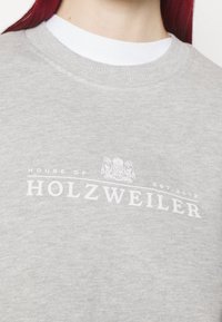 Šedá mikina s kulatým výstřihem vyrobená z měkké tkaniny, na přední straně je bílý potisk s nápisem "HOUSE OF HOLZWEILER" a erbem.