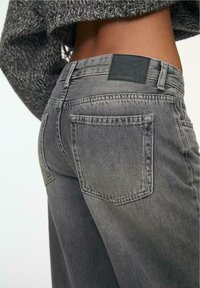 Sive traperice s lagano izbljedjelim završetkom, s dva stražnja džepa, crnom oznakom brenda i detaljem u tonovima od šivanja.