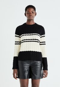 Maglione a trecce nero e crema con motivo a righe orizzontali, caratterizzato da un colletto a girocollo e polsini e orlo a coste. Pantaloni corti in pelle sottostanti.