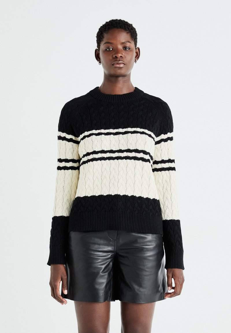 Maglione a trecce nero e crema con motivo a righe orizzontali, caratterizzato da un colletto a girocollo e polsini e orlo a coste. Pantaloni corti in pelle sottostanti.