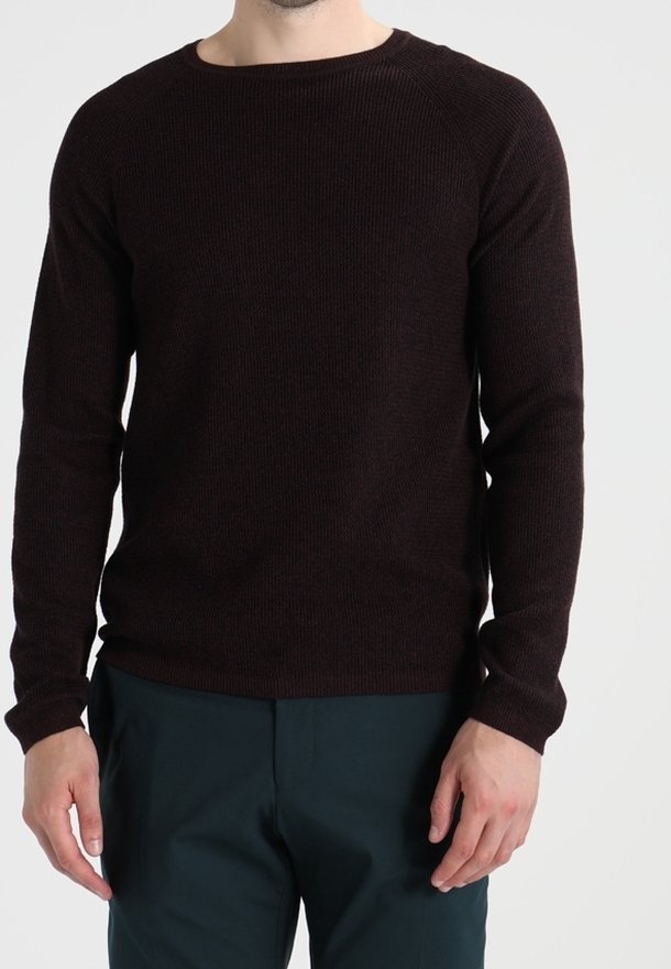 Mężczyzna w dopasowanym, ciemno-brązowym swetrze z długim rękawem i ciemnozielonych spodniach, stojący na jednolitym tle.