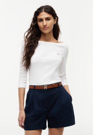 Mujer con cabello largo y oscuro, que lleva una blusa blanca de manga tres cuartos, pantalones cortos azul marino y un cinturón marrón con una hebilla dorada.