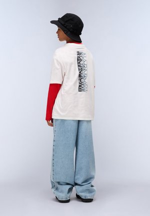 Light beige t-shirt featuring a graphic on the back, layered over a red long-sleeve shirt. Wide blue jeans and a black bucket hat complete the look.
