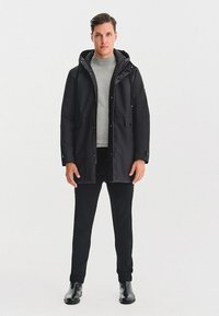 Czarna ocieplana parka z kapturem, przednimi kieszeniami i prostym krojem, noszona na szarym swetrze i czarnych spodniach. Lśniące botki za kostkę.