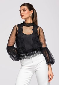 Femme aux longs cheveux attachés en queue de cheval, portant un chemisier en dentelle noire transparente avec des manches bouffantes et un pantalon blanc, debout devant un fond uni.