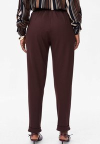 Persoon draagt een donkerbruine, hooggetailleerde broek met taps toelopende pijpen, een gestreept shirt met lange mouwen en paarse schoenen met lage hak, staat met de rug naar voren gericht.