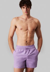Pantaloni da nuoto viola con vita elasticizzata e cordino. Presentano tasche laterali e un tessuto liscio e leggero. Senza fantasie o dettagli.