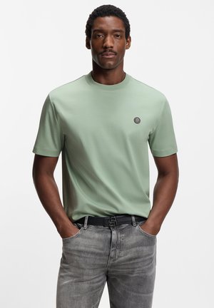 Hellgrünes T-Shirt mit kurzen Ärmeln und rundem Ausschnitt, versehen mit einem kleinen Logo auf der Brust, kombiniert mit dunkelgrauen Denim-Jeans und einem schwarzen Gürtel.